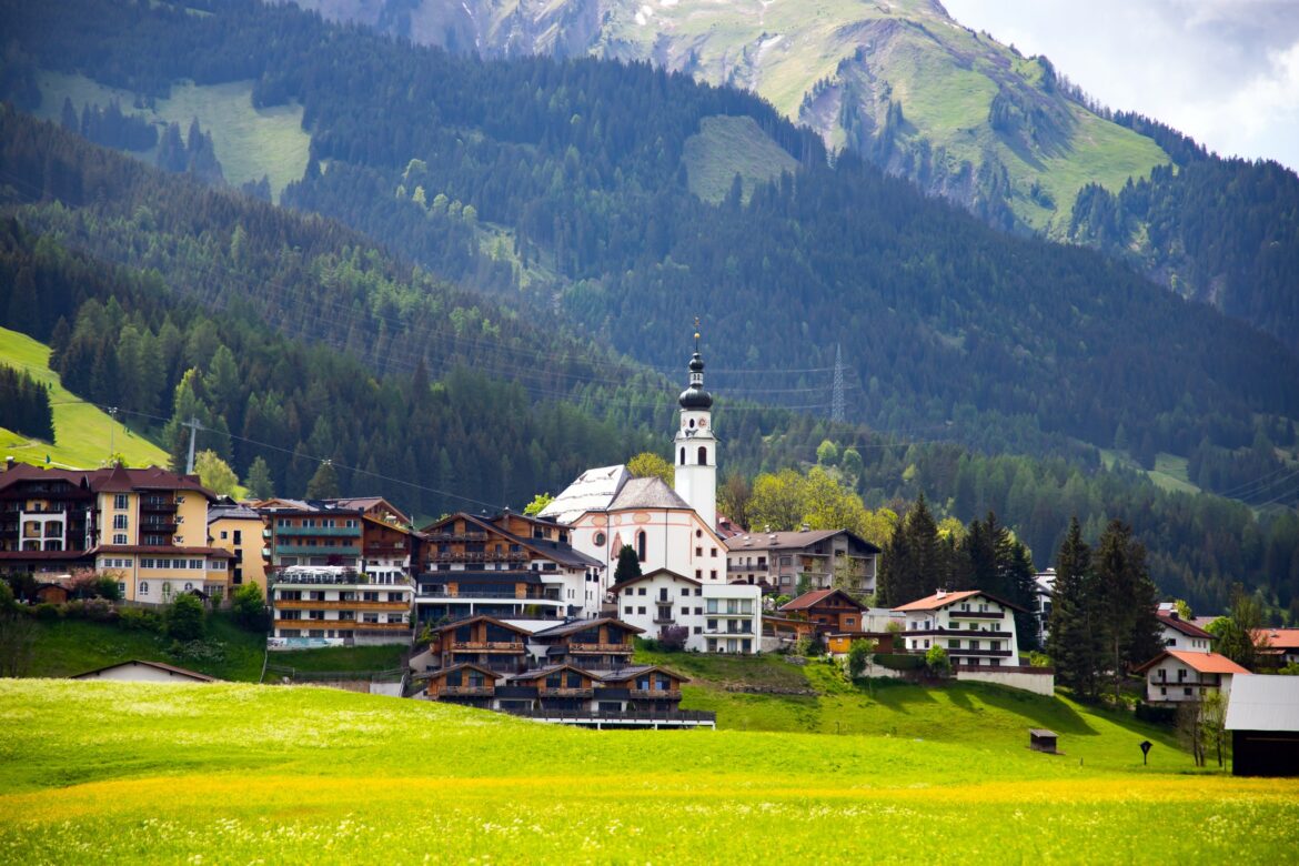 Alpendorf mit weißer Kirche und Hotels am bewaldeten Berghang, davor eine blühende Wiese